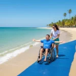Pessoa em cadeira de rodas adaptada na beira da água em uma praia paradisíaca de Maceió, com infraestrutura de acesso e palmeiras, destacando as praias acessíveis em Maceió para cadeirantes.